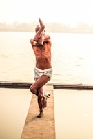A person is performing a yoga pose near a body of water, standing on one leg with arms intertwined. The setting is serene, with a calm water surface and muted background details.
