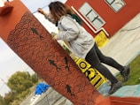 A person wearing a grey hoodie and black jeans is painting a column with intricate black wave patterns and fish designs over a reddish-orange base. The area appears to be an outdoor space with a red building in the background adorned with large, abstract framed paintings. The ground is a mix of concrete and grass.