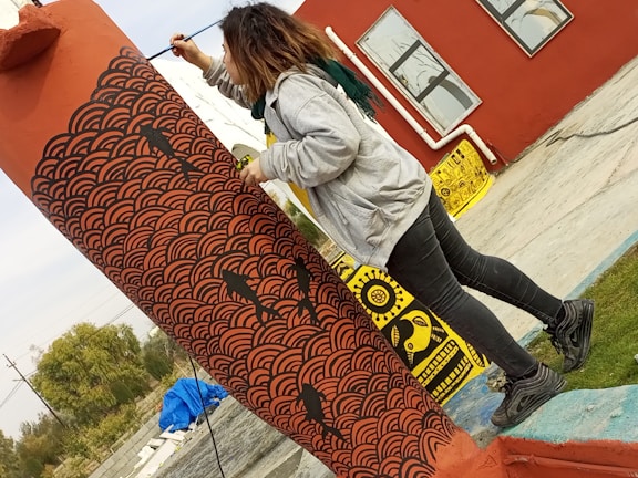A person wearing a grey hoodie and black jeans is painting a column with intricate black wave patterns and fish designs over a reddish-orange base. The area appears to be an outdoor space with a red building in the background adorned with large, abstract framed paintings. The ground is a mix of concrete and grass.
