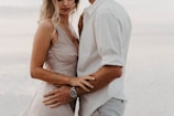 A couple standing closely together, partially visible, showcasing intimate body language. The woman wears a light-colored dress, and the man is in a white shirt. There is a soft and serene background, likely a beach setting.