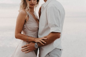 A couple standing closely together, partially visible, showcasing intimate body language. The woman wears a light-colored dress, and the man is in a white shirt. There is a soft and serene background, likely a beach setting.