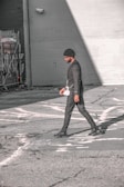 A person walks across an urban street, dressed in a stylish black outfit and a beanie. The scene features urban elements like graffiti and security cameras on the surrounding walls, with strong sunlight creating shadows on the pavement.