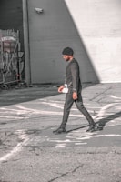 A person walks across an urban street, dressed in a stylish black outfit and a beanie. The scene features urban elements like graffiti and security cameras on the surrounding walls, with strong sunlight creating shadows on the pavement.