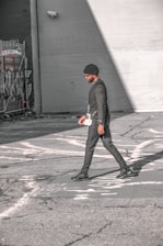 A person walks across an urban street, dressed in a stylish black outfit and a beanie. The scene features urban elements like graffiti and security cameras on the surrounding walls, with strong sunlight creating shadows on the pavement.