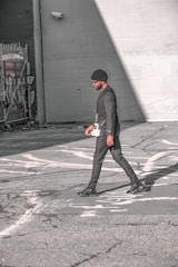 A person walks across an urban street, dressed in a stylish black outfit and a beanie. The scene features urban elements like graffiti and security cameras on the surrounding walls, with strong sunlight creating shadows on the pavement.