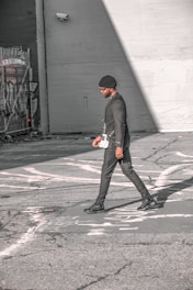 A person walks across an urban street, dressed in a stylish black outfit and a beanie. The scene features urban elements like graffiti and security cameras on the surrounding walls, with strong sunlight creating shadows on the pavement.