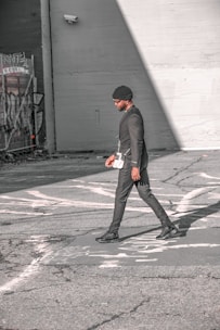A person walks across an urban street, dressed in a stylish black outfit and a beanie. The scene features urban elements like graffiti and security cameras on the surrounding walls, with strong sunlight creating shadows on the pavement.
