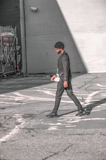 A person walks across an urban street, dressed in a stylish black outfit and a beanie. The scene features urban elements like graffiti and security cameras on the surrounding walls, with strong sunlight creating shadows on the pavement.
