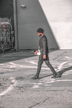 A person walks across an urban street, dressed in a stylish black outfit and a beanie. The scene features urban elements like graffiti and security cameras on the surrounding walls, with strong sunlight creating shadows on the pavement.