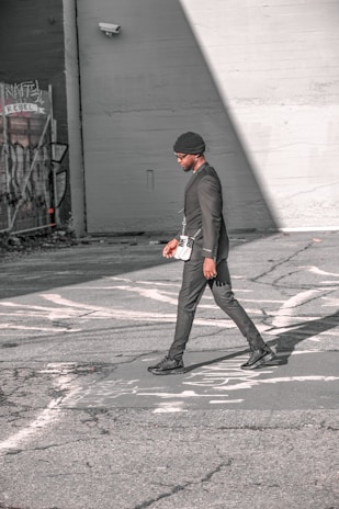 A person walks across an urban street, dressed in a stylish black outfit and a beanie. The scene features urban elements like graffiti and security cameras on the surrounding walls, with strong sunlight creating shadows on the pavement.