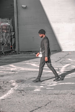 A person walks across an urban street, dressed in a stylish black outfit and a beanie. The scene features urban elements like graffiti and security cameras on the surrounding walls, with strong sunlight creating shadows on the pavement.