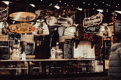 A festive market stall is adorned with warm, inviting lights and decorative signs indicating various holiday beverages. There is a snowman figurine alongside bottles and glasses on display. The overall vibe is cozy and lively, with a notable emphasis on the holiday spirit.