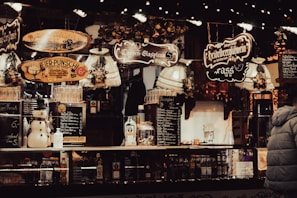 A festive market stall is adorned with warm, inviting lights and decorative signs indicating various holiday beverages. There is a snowman figurine alongside bottles and glasses on display. The overall vibe is cozy and lively, with a notable emphasis on the holiday spirit.