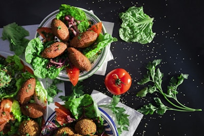 A vibrant assortment of dishes featuring kibbeh, a Middle Eastern dish made of bulgur, minced onions, and ground lean beef. The kibbeh is garnished with fresh lettuce, parsley, sliced tomatoes, and other greens. A whole tomato and fresh herbs are scattered around, enhancing the table setting.