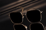 A pair of sunglasses with a sleek, square frame featuring reflective lenses. The sunglasses are set against a dark and shiny surface, creating a mirrored effect. Subtle lighting casts soft shadows adding to the sophisticated appearance.