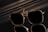 A pair of sunglasses with a sleek, square frame featuring reflective lenses. The sunglasses are set against a dark and shiny surface, creating a mirrored effect. Subtle lighting casts soft shadows adding to the sophisticated appearance.