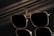 A pair of sunglasses with a sleek, square frame featuring reflective lenses. The sunglasses are set against a dark and shiny surface, creating a mirrored effect. Subtle lighting casts soft shadows adding to the sophisticated appearance.