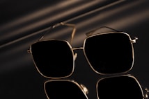 A pair of sunglasses with a sleek, square frame featuring reflective lenses. The sunglasses are set against a dark and shiny surface, creating a mirrored effect. Subtle lighting casts soft shadows adding to the sophisticated appearance.