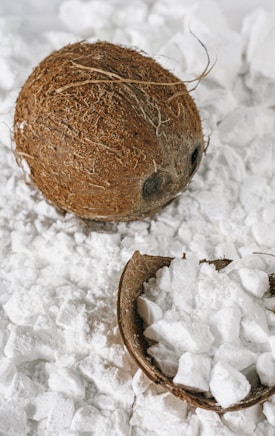 A whole coconut and a broken coconut shell are surrounded by white, powdery chunks resembling coconut flakes or coconut-related product. The background is filled with similar white substances.