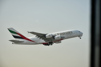 An Airbus A380 airplane bearing the Emirates airline branding is in mid-flight against a clear sky. The aircraft's engines and landing gear are visible.