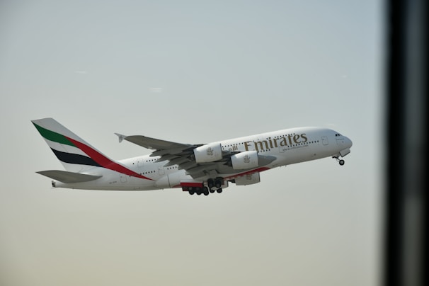 An Airbus A380 airplane bearing the Emirates airline branding is in mid-flight against a clear sky. The aircraft's engines and landing gear are visible.