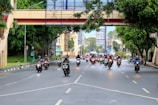 A group of motorcyclists travels along a wide urban road lined with trees and buildings. An overhead pedestrian bridge crosses the street, and the scene is lively with traffic. The motorcyclists are wearing helmets, some with jackets or rain gear.