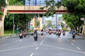 A group of motorcyclists travels along a wide urban road lined with trees and buildings. An overhead pedestrian bridge crosses the street, and the scene is lively with traffic. The motorcyclists are wearing helmets, some with jackets or rain gear.