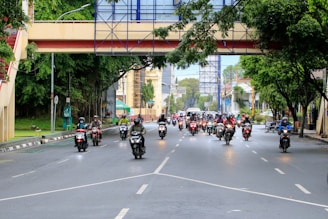 A group of motorcyclists travels along a wide urban road lined with trees and buildings. An overhead pedestrian bridge crosses the street, and the scene is lively with traffic. The motorcyclists are wearing helmets, some with jackets or rain gear.