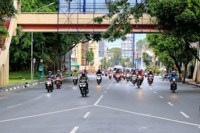 A group of motorcyclists travels along a wide urban road lined with trees and buildings. An overhead pedestrian bridge crosses the street, and the scene is lively with traffic. The motorcyclists are wearing helmets, some with jackets or rain gear.