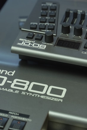 Close-up view of parts of Roland synthesizer models JD-08 and JD-800. The image features the control buttons, knobs, and sliders of the instruments along with the branding and model names on the panels.