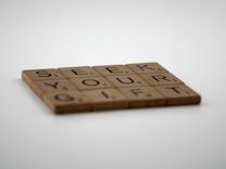 Wooden letter tiles arranged to form the words 'SEEK YOUR GIFT'. The tiles are placed on a white surface, with focus on the letters while the background appears soft and blurred.