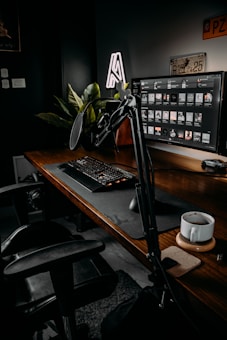A well-organized workspace featuring a large monitor displaying a software interface, a mechanical keyboard, and a professional microphone setup on a wooden desk. A coffee mug sits on a coaster, and there are lush green plants in the background. The space is dimly lit, creating a cozy and focused atmosphere.