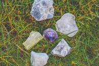 A collection of rough gemstones lying on a patch of grass. The stones vary in color, including clear, purple, and pale yellow, and have a raw, unpolished appearance.