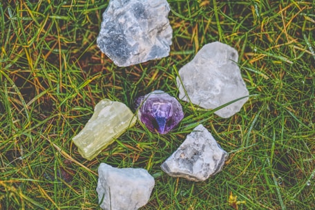 A collection of rough gemstones lying on a patch of grass. The stones vary in color, including clear, purple, and pale yellow, and have a raw, unpolished appearance.