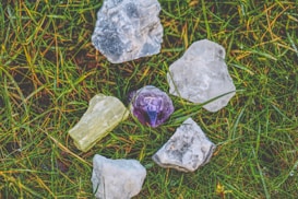 A collection of rough gemstones lying on a patch of grass. The stones vary in color, including clear, purple, and pale yellow, and have a raw, unpolished appearance.