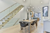 A modern dining and kitchen area with minimalist design. The room features a wooden dining table surrounded by sleek black chairs. To the side, there's a staircase with glass railing leading to the upper floor. The kitchen is equipped with contemporary appliances and stylish pendant lights. Large windows allow ample natural light to flood the space, while wooden flooring adds warmth to the room.