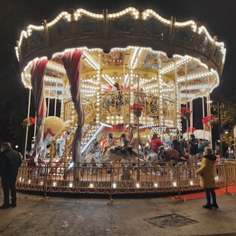 A brightly lit carousel with ornate decorations spinning at night. Children and adults are riding on various animals like horses and zebras. The structure is adorned with numerous lights, flowers, and red ribbons, creating a festive and joyful atmosphere.