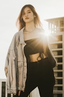 A person stands confidently in a fashionable outfit, featuring a denim jacket draped over a black crop top and dark pants. Sunlight shines from behind, creating a backlighting effect and highlighting the person's hair. A modern building is partially visible in the background, adding an urban touch.