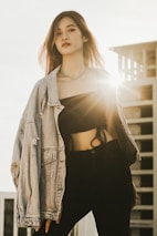 A person stands confidently in a fashionable outfit, featuring a denim jacket draped over a black crop top and dark pants. Sunlight shines from behind, creating a backlighting effect and highlighting the person's hair. A modern building is partially visible in the background, adding an urban touch.