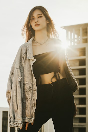 A person stands confidently in a fashionable outfit, featuring a denim jacket draped over a black crop top and dark pants. Sunlight shines from behind, creating a backlighting effect and highlighting the person's hair. A modern building is partially visible in the background, adding an urban touch.