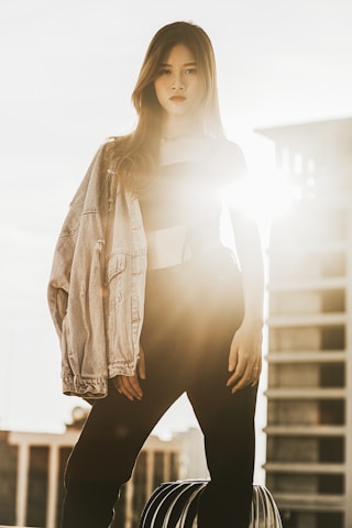 A young woman stands confidently on a rooftop, backlit by the sun, which creates a halo effect and a distinct lens flare. She's wearing a denim jacket draped over one shoulder and dark pants. High-rise buildings are slightly blurred in the background, emphasizing the urban setting.