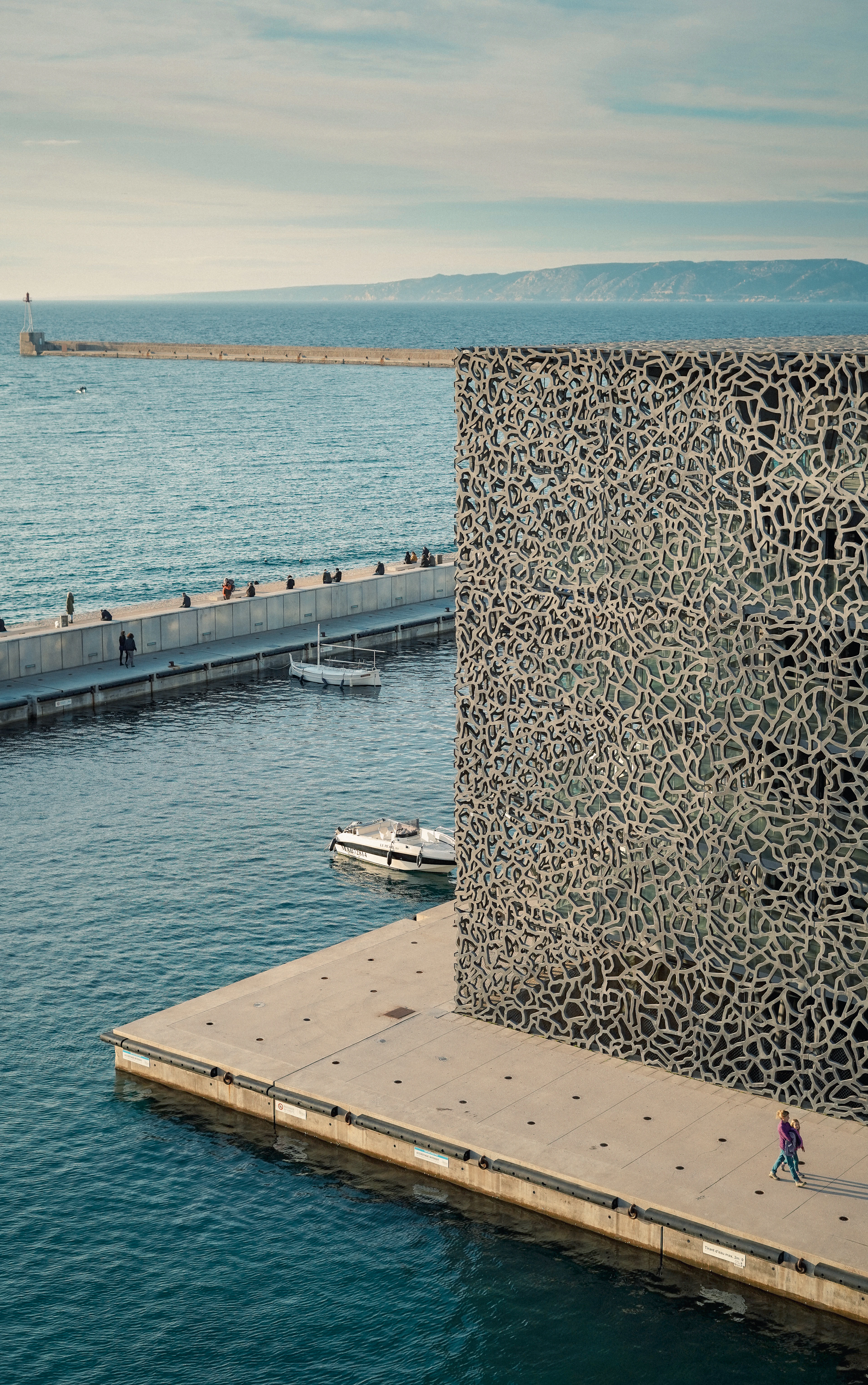 The MuCEM in Marseille from above, its dark filigree concrete lattice standing on a bare quay over green Mediterranean water, walkers strung along the footbridge and the breakwater and hills of the Cote Bleue beyond.