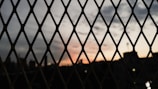 A metal fence with diamond-shaped openings is in the foreground, silhouetted against a skyline during sunset. The sky is a gradient of soft colors transitioning from orange to gray, with scattered clouds visible.