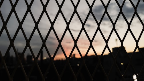 A metal fence with diamond-shaped openings is in the foreground, silhouetted against a skyline during sunset. The sky is a gradient of soft colors transitioning from orange to gray, with scattered clouds visible.