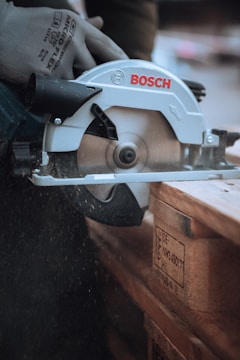 A circular saw is being operated by a person wearing protective gloves. The saw is actively cutting through a wooden pallet, and sawdust is visibly dispersing into the air. The brand name on the saw is Bosch.
