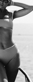 A person in a swimsuit is posing by the water, with a relaxed and confident posture. They are wearing sunglasses and a navel piercing is visible. The image has a black and white filter, adding a classic touch.