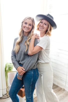Two people stand close together, smiling warmly. One person wears a grey sweater and jeans, while the other wears a white shirt and pants, accessorizing with a wide-brimmed hat. The background is softly lit with bright natural light coming in from a window, providing a cozy and inviting atmosphere.