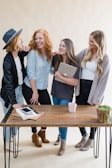 Four women are standing together, laughing and smiling, suggesting a joyful and friendly atmosphere. They are gathered around a wooden table, with one woman holding a laptop. A potted plant and some notebooks are also visible on the table.