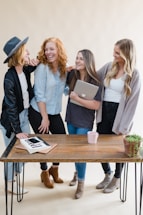 Four women are standing together, laughing and smiling, suggesting a joyful and friendly atmosphere. They are gathered around a wooden table, with one woman holding a laptop. A potted plant and some notebooks are also visible on the table.