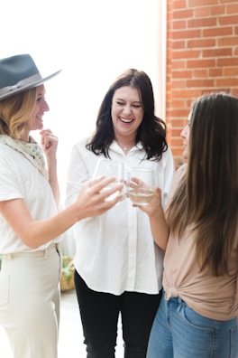 Three women are gathered together, smiling and laughing while holding drinks. They appear to be enjoying a social moment in a bright space with natural light and a brick wall in the background.